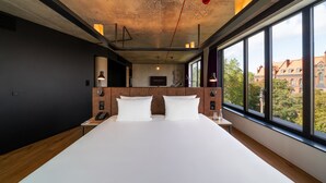 A modern hotel room with a large bed, a wooden headboard, and a view of the city through large windows. - The Bridge Wroclaw - MGallery (Wroclaw)