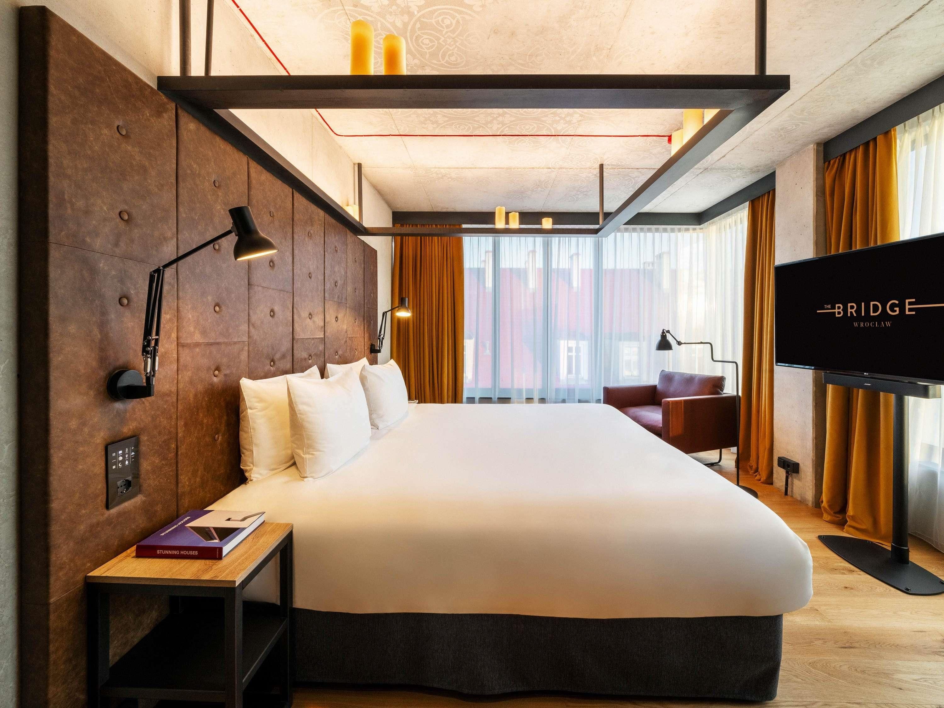 A modern hotel room with a large bed, a TV, a desk, and a view of buildings through large windows.