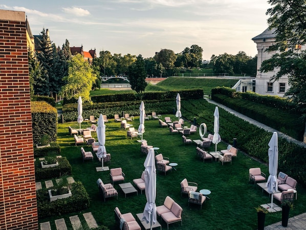 An outdoor seating area with chairs and tables, surrounded by greenery and a building in the background. - The Bridge Wroclaw - MGallery (Wroclaw)
