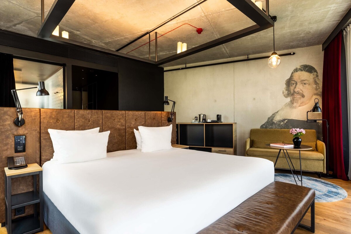 A modern hotel room with a large bed, a sofa, a coffee table, a desk, and a wall-mounted TV.
