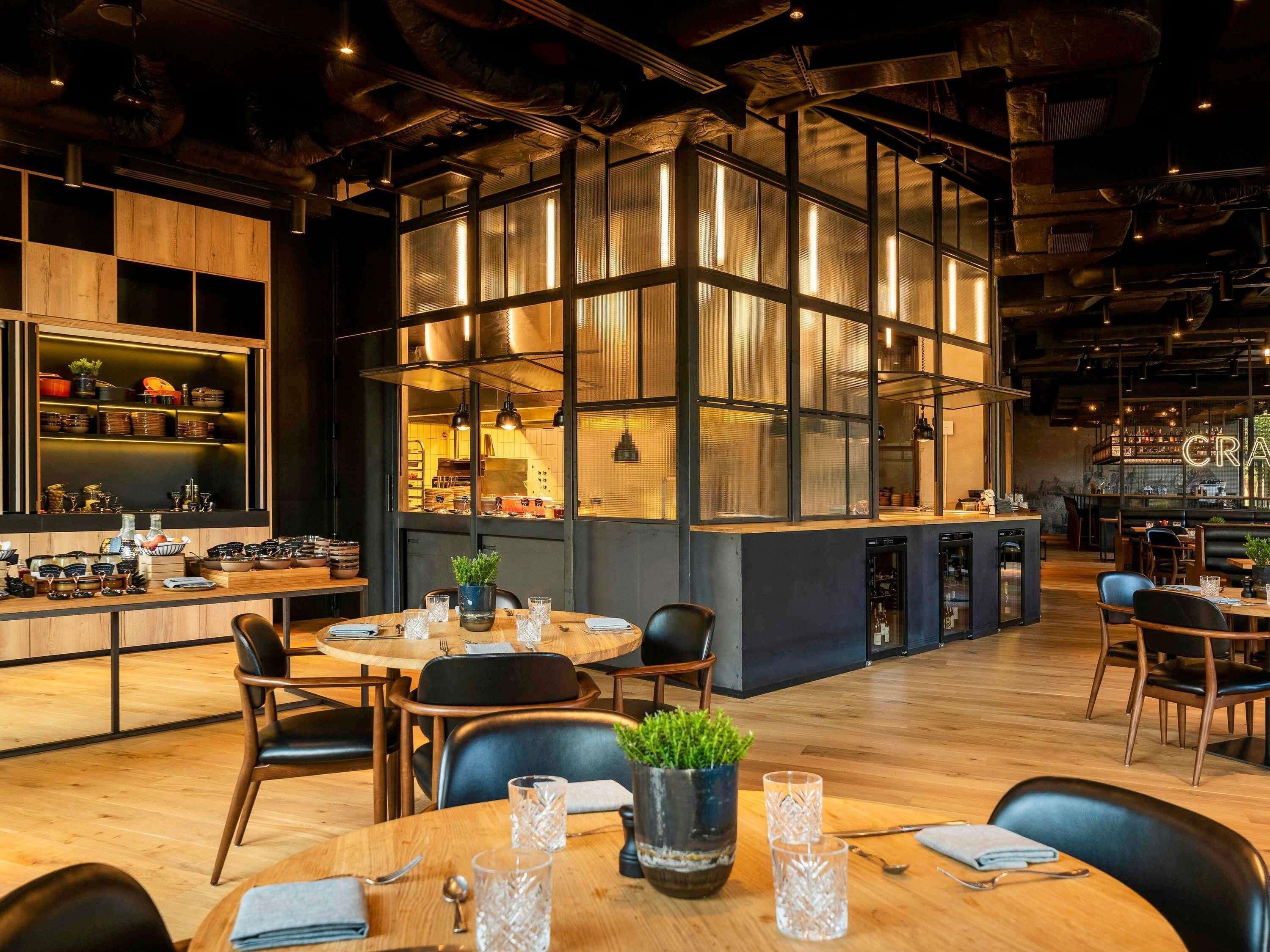 Un interior de restaurante moderno con mesas de madera, sillas negras y un gran ventanal con paneles iluminados.