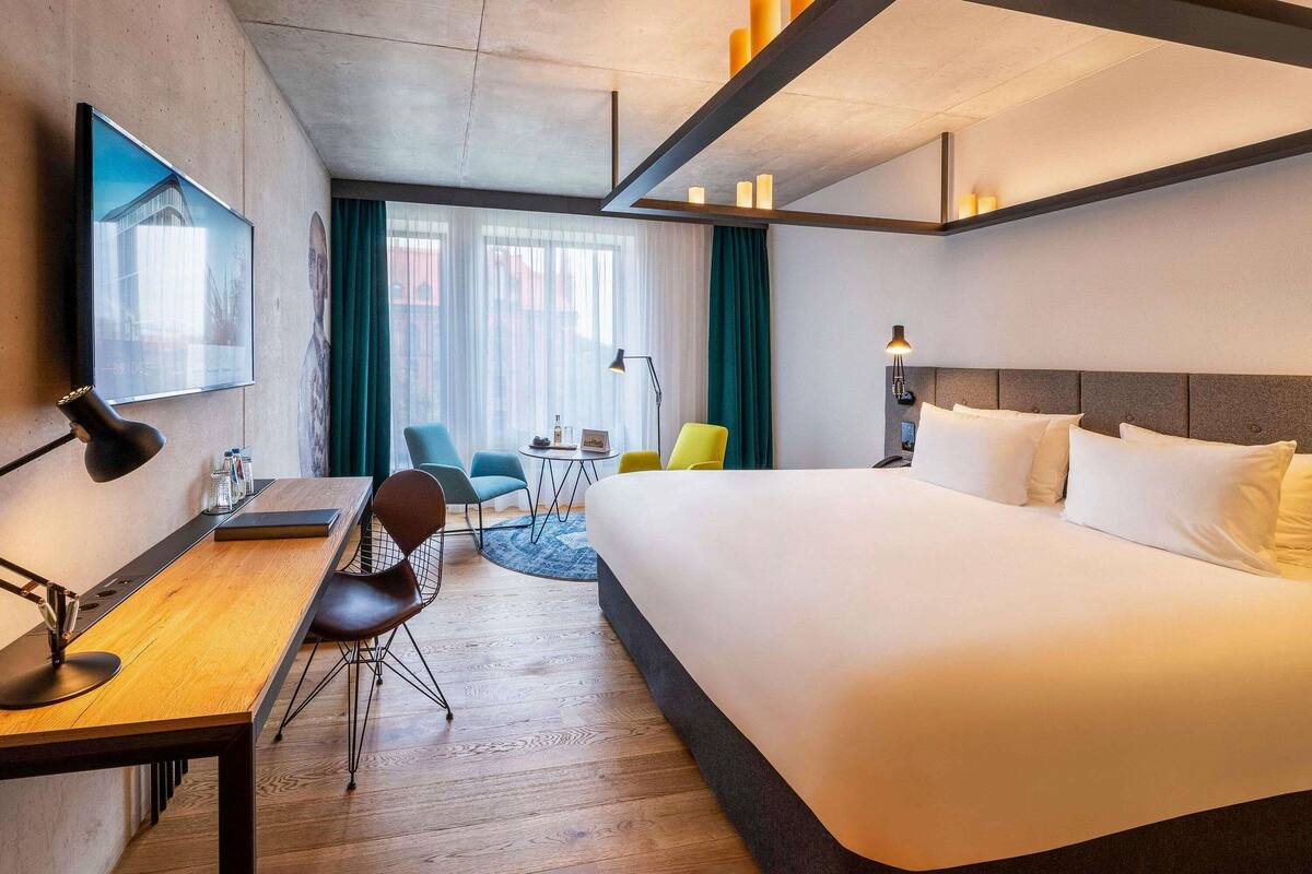 A modern hotel room with a large bed, a desk with a chair, a TV, and a view of the city through large windows.