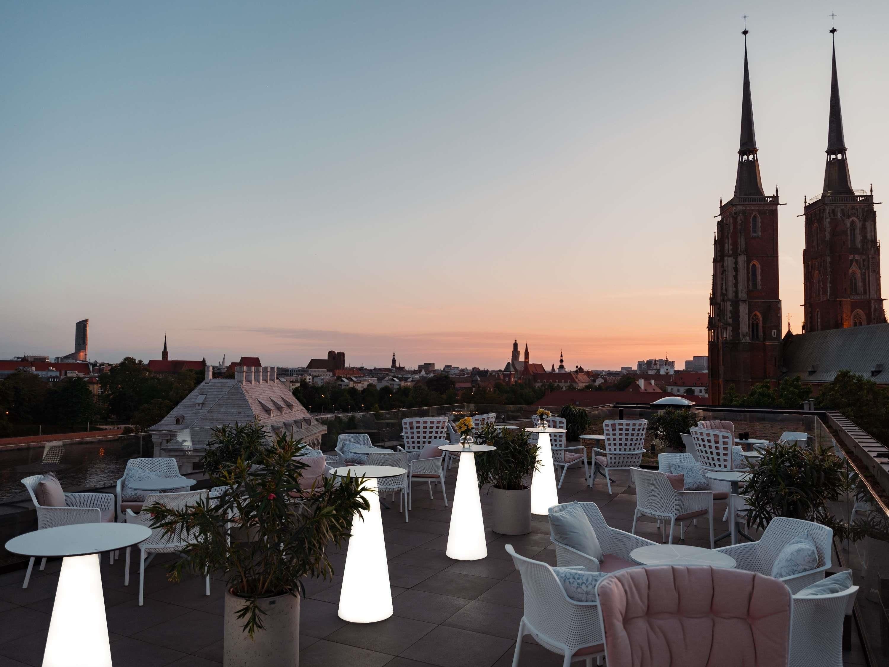 A rooftop terrace with outdoor seating, potted plants, and a view of a cityscape including a church with twin spires.
