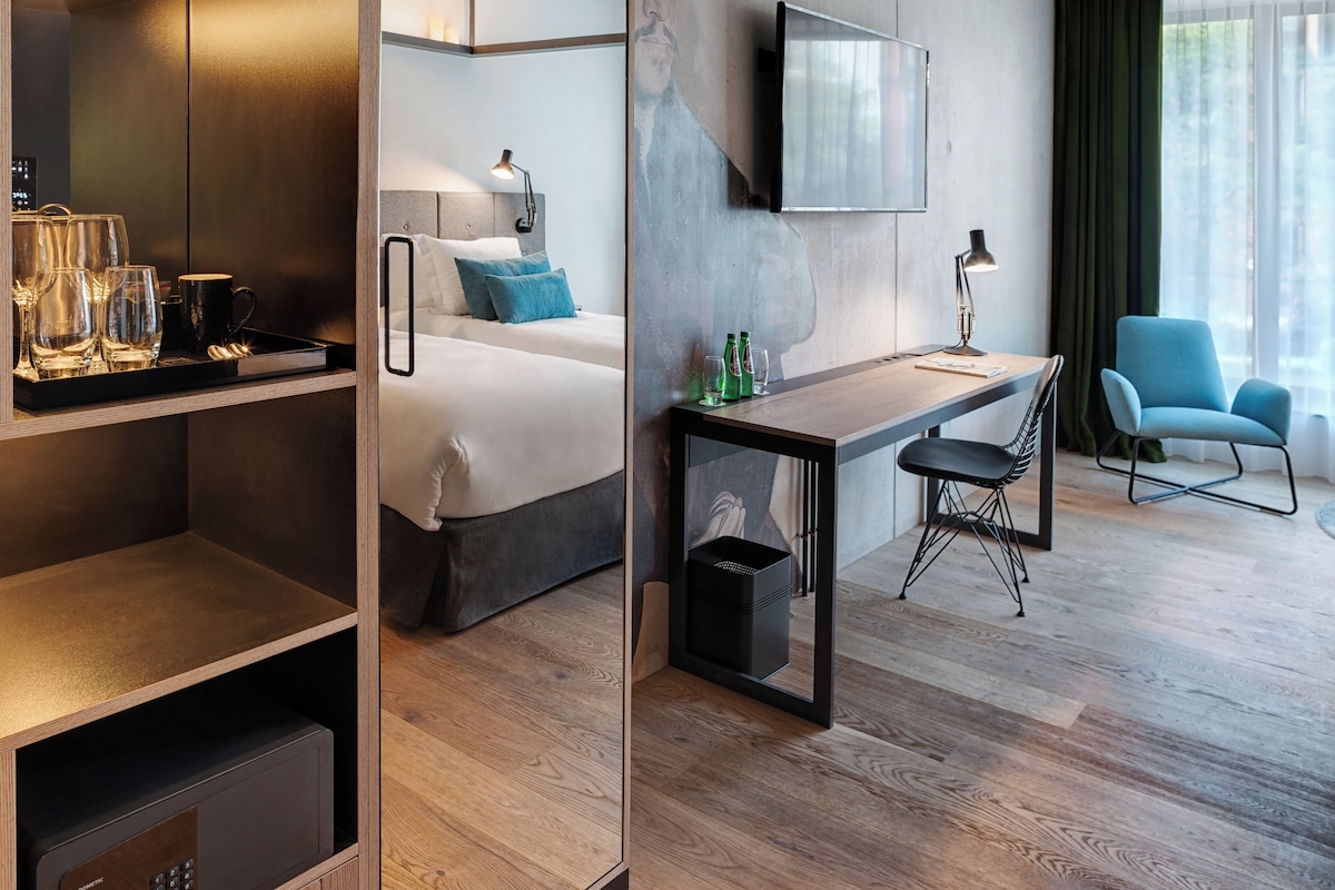 A modern hotel room with a bed, desk, chair, TV, and a view of the outdoors.