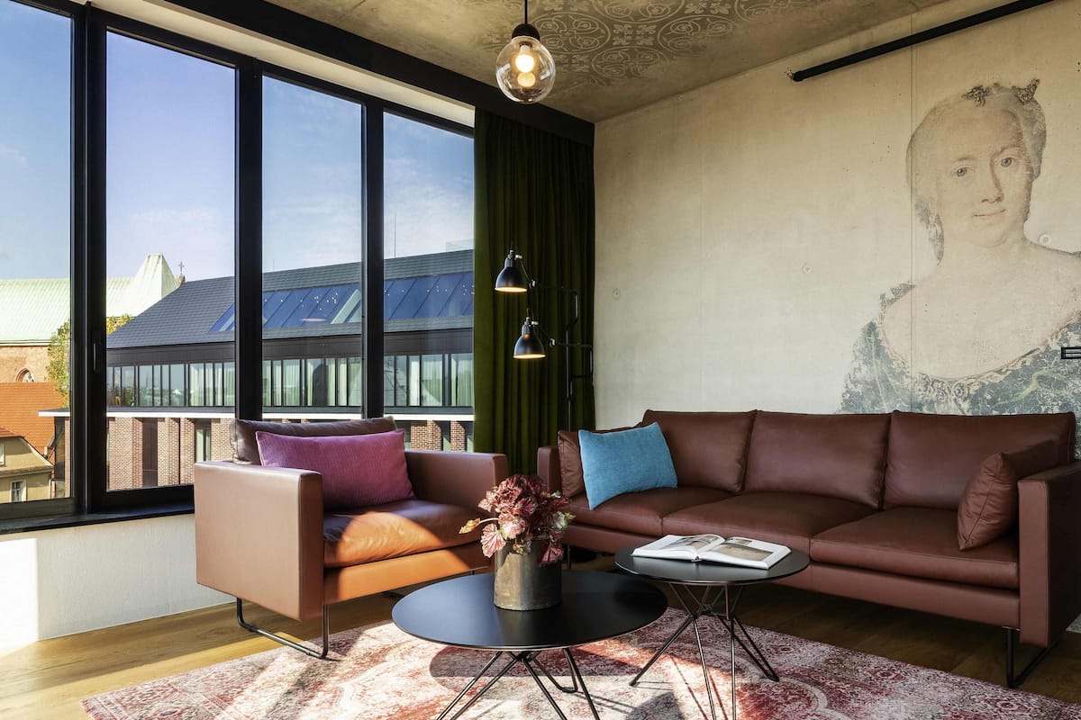 A modern living room with a large window, a brown leather sofa, a round coffee table, and a green curtain.