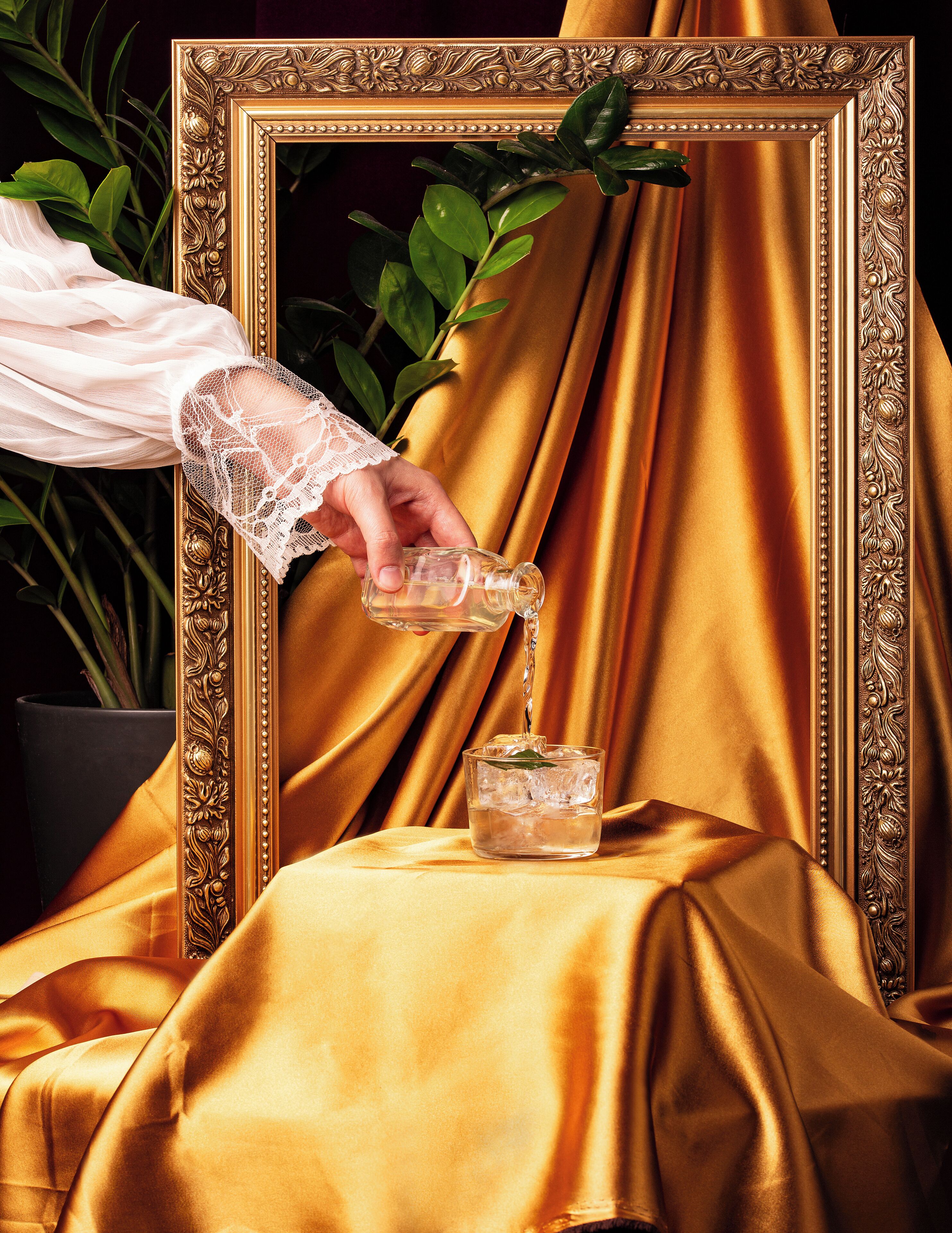 A hand pouring a clear liquid into a glass with ice cubes, set against a golden fabric backdrop and an ornate frame.