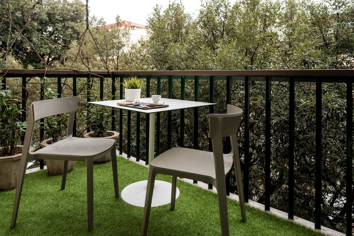 A small outdoor seating area with two chairs and a table, surrounded by a green railing and lush foliage.