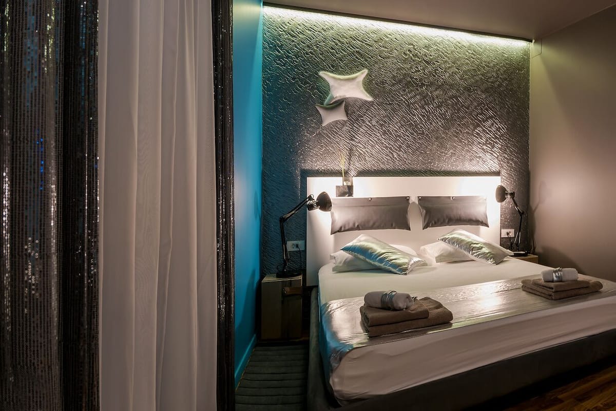 A modern hotel room with a bed, bedside lamps, a nightstand, and a bathroom visible through an open door.