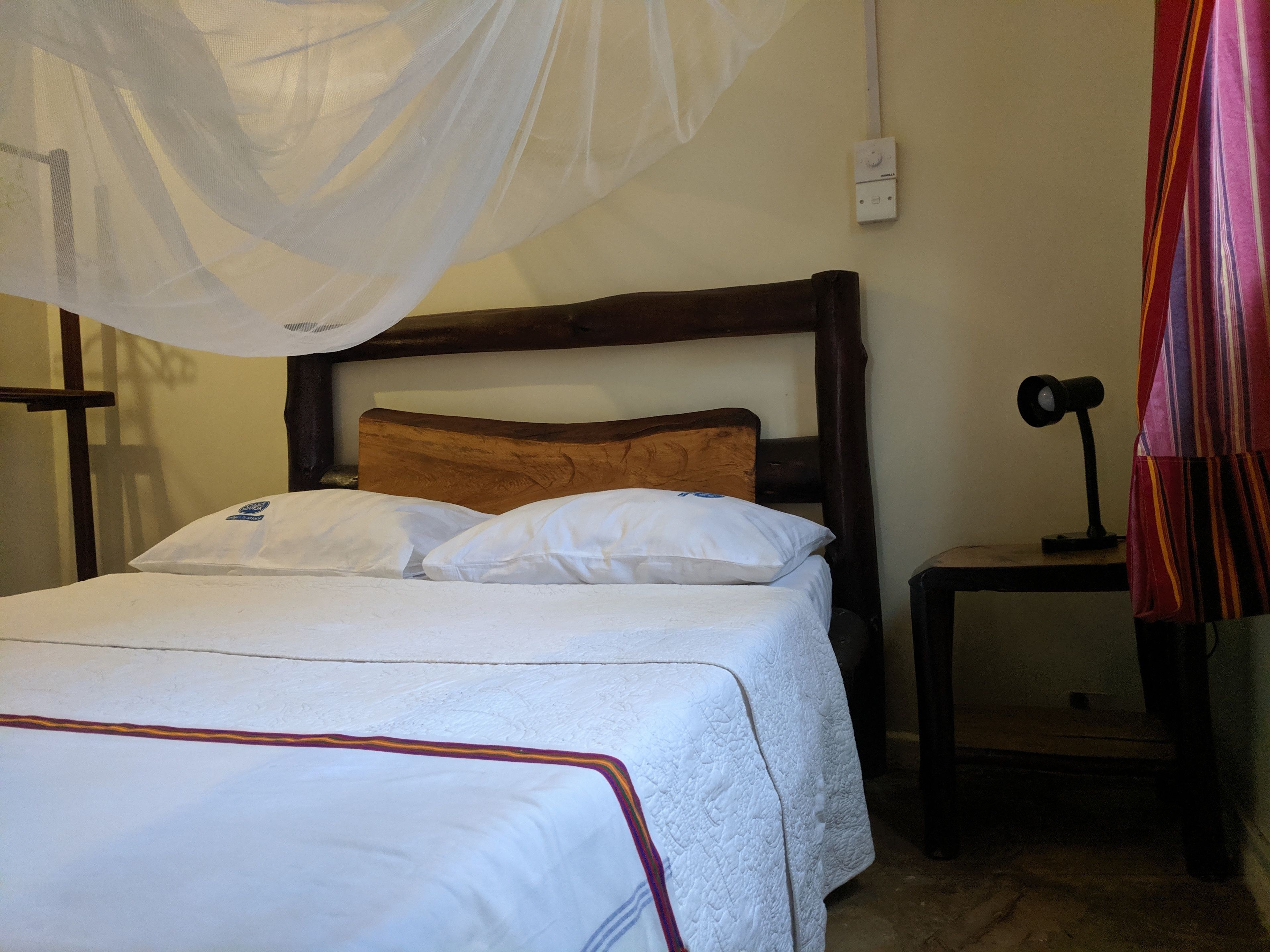 Una cama con ropa de cama blanca y un cabecero de madera, una mesita de noche con una lámpara y una mosquitera sobre la cama.