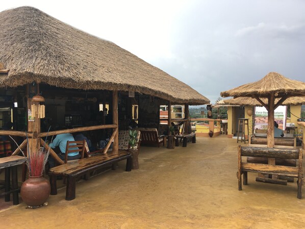 Un espacio de estar al aire libre rústico con estructuras de techo de paja, bancos de madera y una planta grande en maceta.