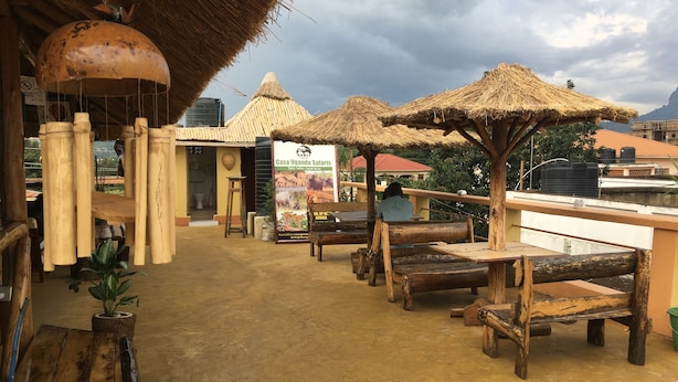Un espace extérieur rustique aménagé avec des parasols en chaume, des bancs en bois et un panneau indiquant « Casa Uganda Safaris ».