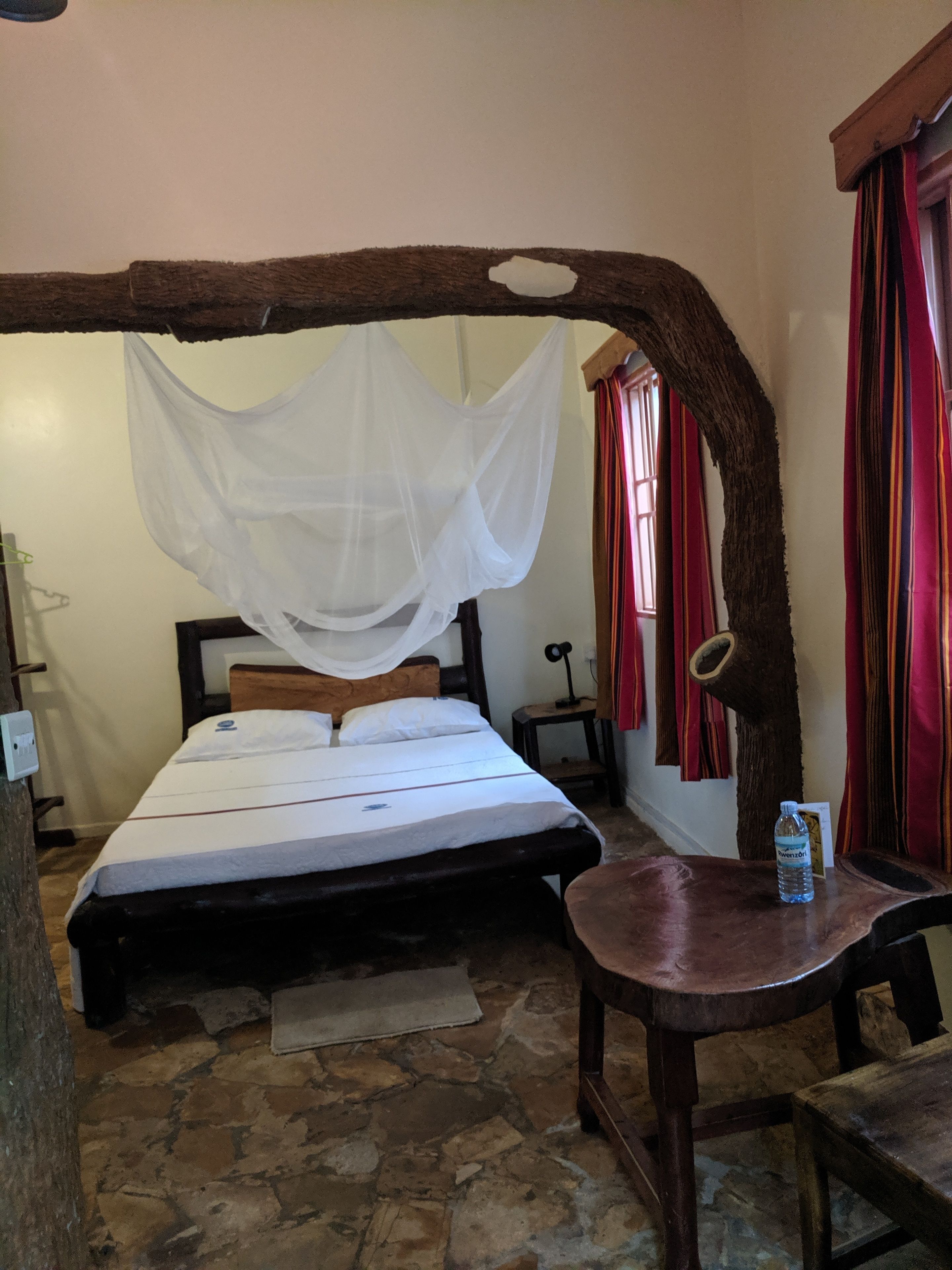 A room with a bed under a canopy, a wooden table with a bottle, and a stone floor.