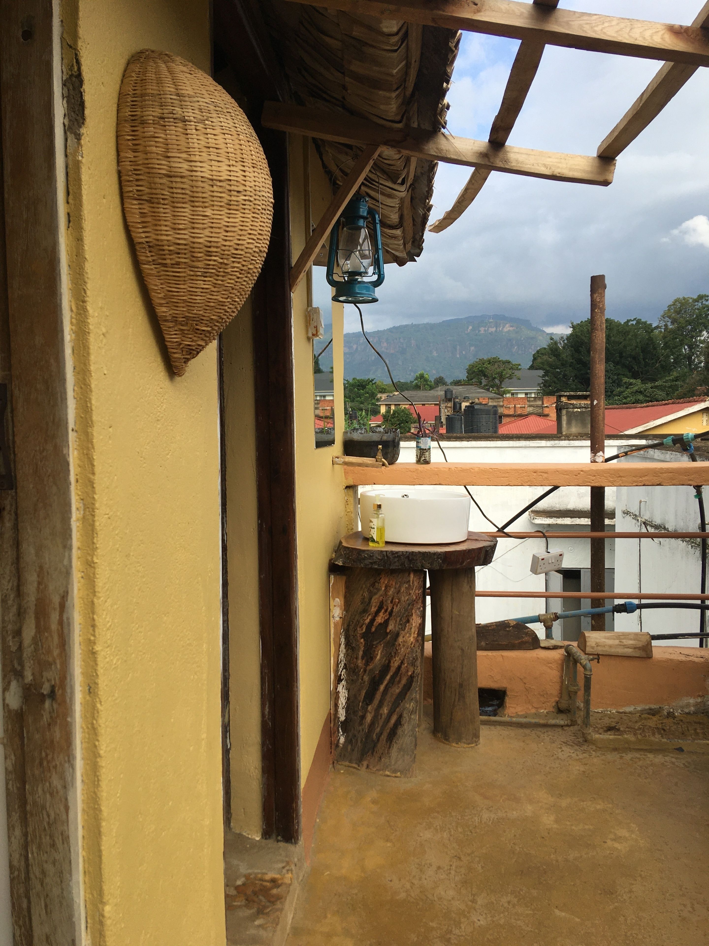 A rustic bathroom with a yellow wall, a woven basket, a blue lantern, and a view of a cityscape with mountains in the background.