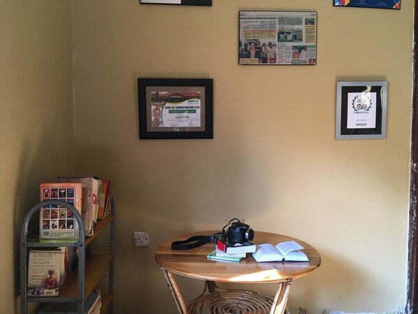 A room with a table holding books and a camera, a shelf with books, and framed certificates on the wall. - Casa Del Turista (Mbale)