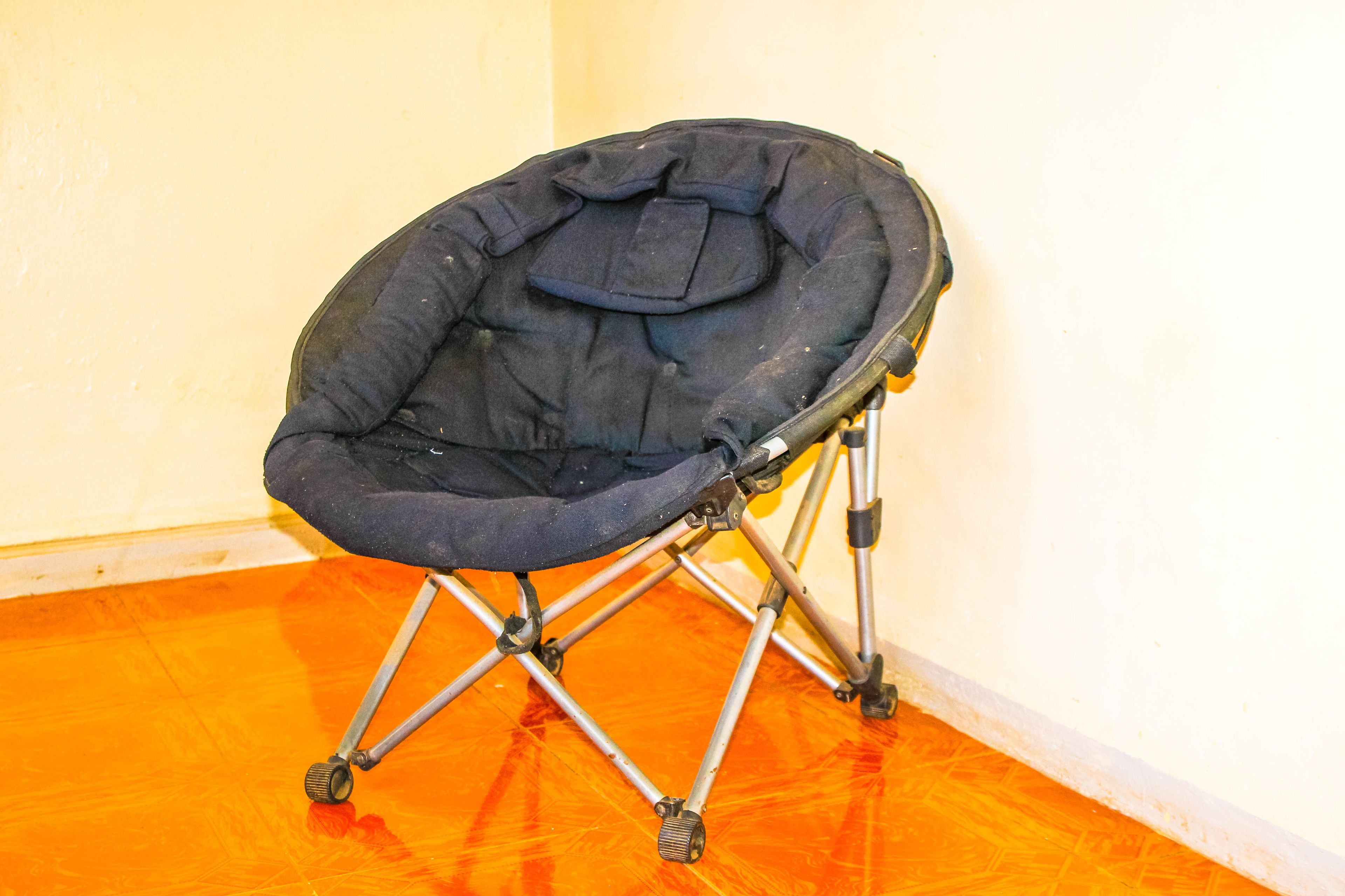 A black camping chair with a cushion, metal frame, and wheels, placed on an orange floor against a white wall.
