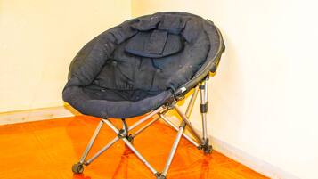 Una silla plegable para acampar de color negro con cojín, estructura metálica y ruedas, ubicada sobre un suelo naranja frente a una pared blanca.