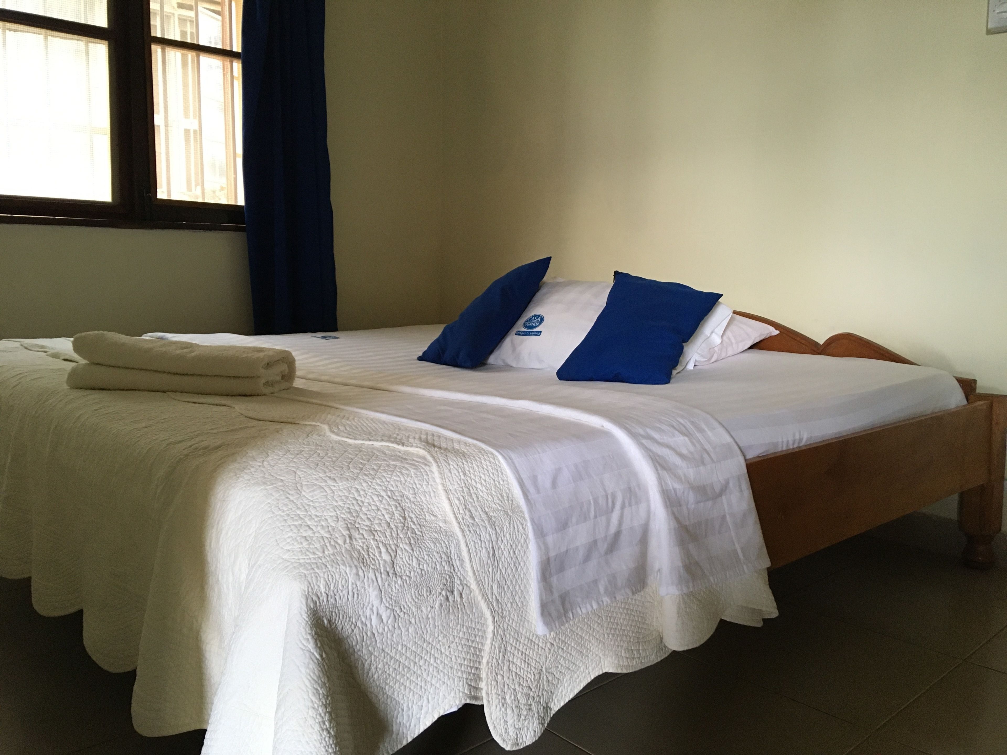 Una cama bien hecha con sábanas blancas y almohadas azules, una toalla doblada y una ventana con cortinas.