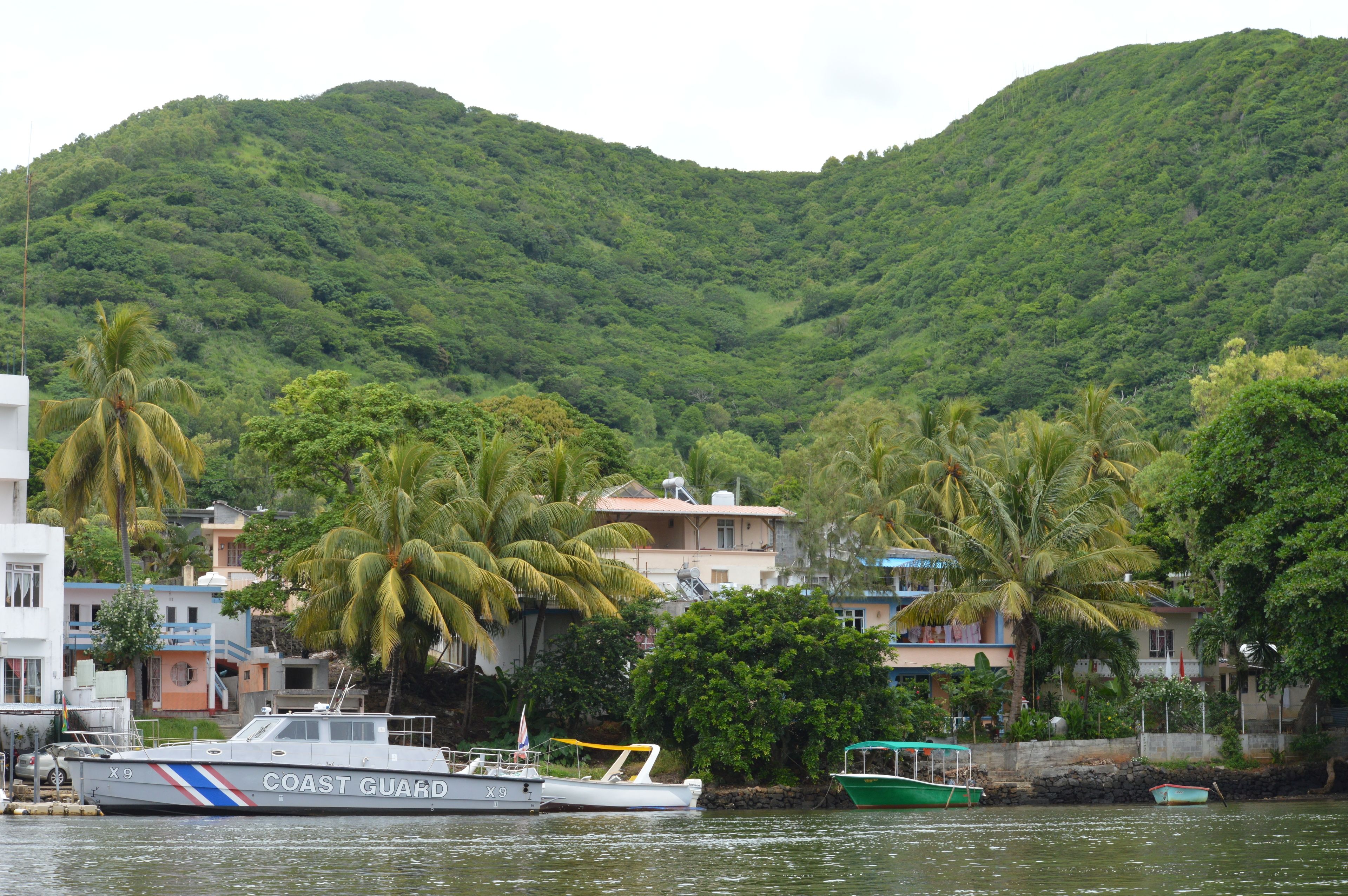 Uma área costeira com um barco da Guarda Costeira atracado perto de edifícios e palmeiras, com uma montanha ao fundo.