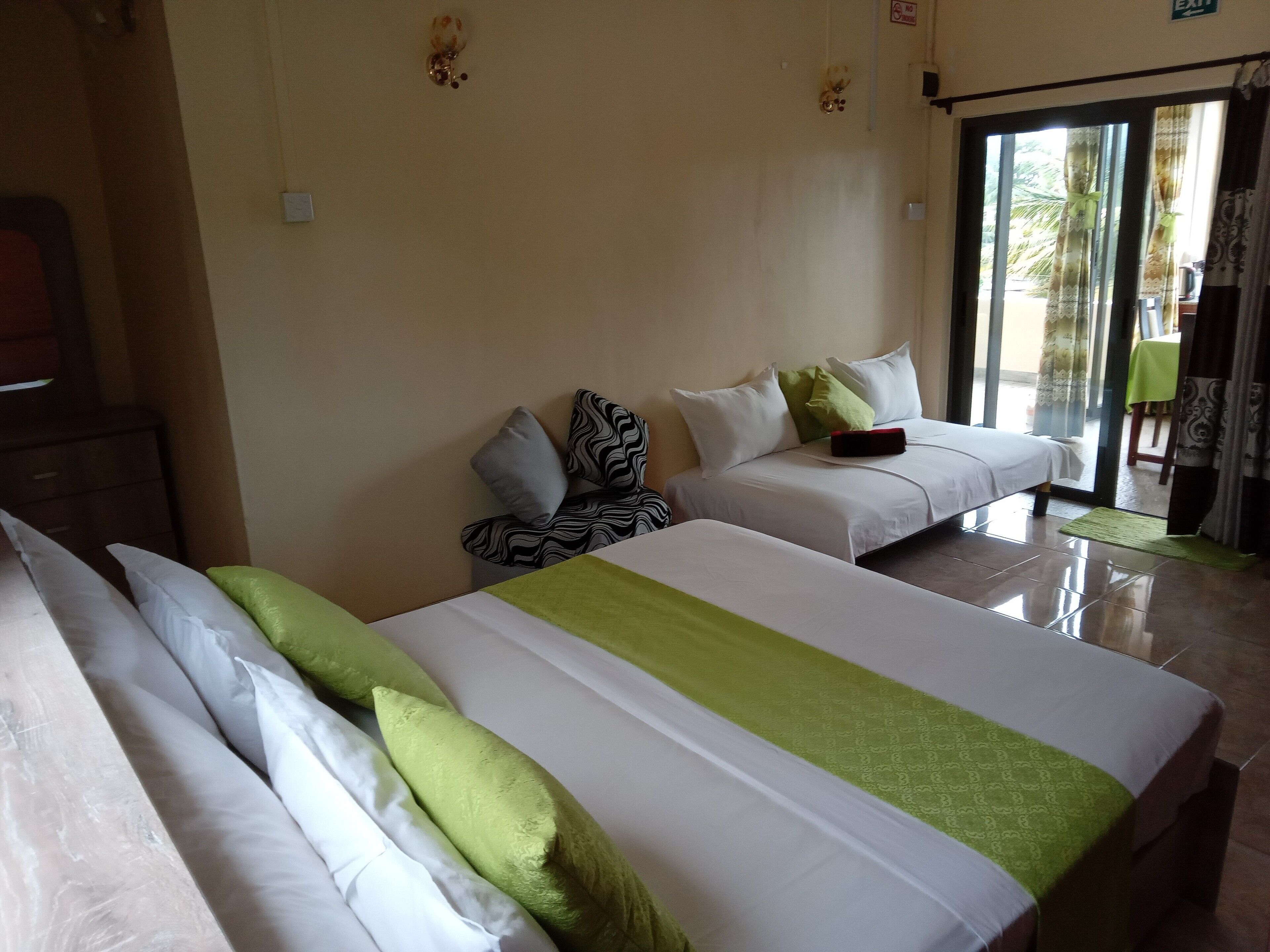 Une chambre d’hôtel avec deux lits, chacun agrémenté de draps blancs et verts, une petite table sur laquelle se trouve un objet rouge, et une vue sur l’extérieur à travers une porte vitrée.