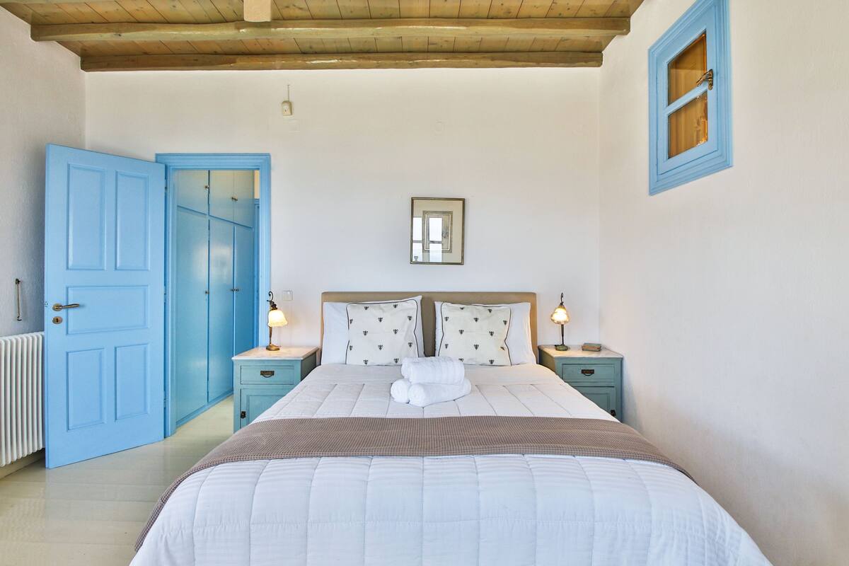 Un dormitorio con una cama grande, dos mesitas de noche con lámparas, una puerta azul y un ventanal pequeño.