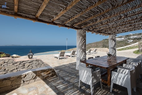 A wooden pergola with a view of the sea, stone walls, and a table with chairs.