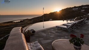 An infinity pool with lounge chairs and a potted plant, overlooking a coastal landscape with a sunset. - Anerousses Mykonian Traditional Houses (Mykonos)