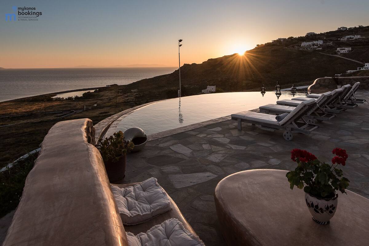 Una piscina infinita con sillones de descanso y una planta en maceta, con vistas a un paisaje costero al atardecer.