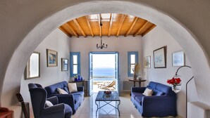 A living room with two blue sofas, a coffee table, and a guitar. There is a view of the beach through an open door. - Anerousses Mykonian Traditional Houses (Mykonos)