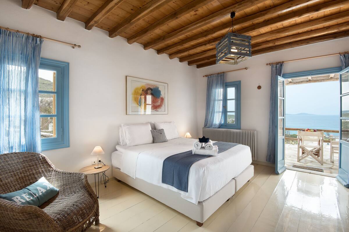 Un dormitorio con una cama, una silla de mimbre, una mesa con lámpara y vistas al exterior.