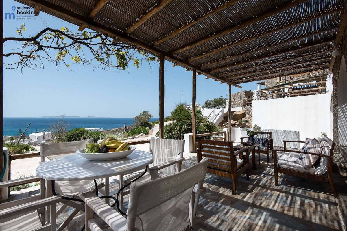 Un espacio de estar techado al aire libre con mesa y sillas, con vistas panorámicas al mar y edificios.