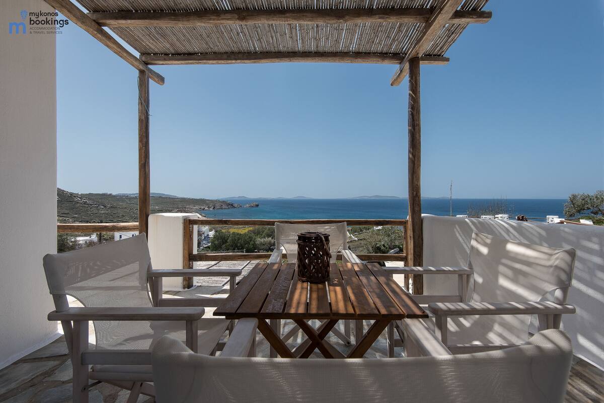 Una mesa de madera y sillas blancas bajo una pérgola con vista al mar.