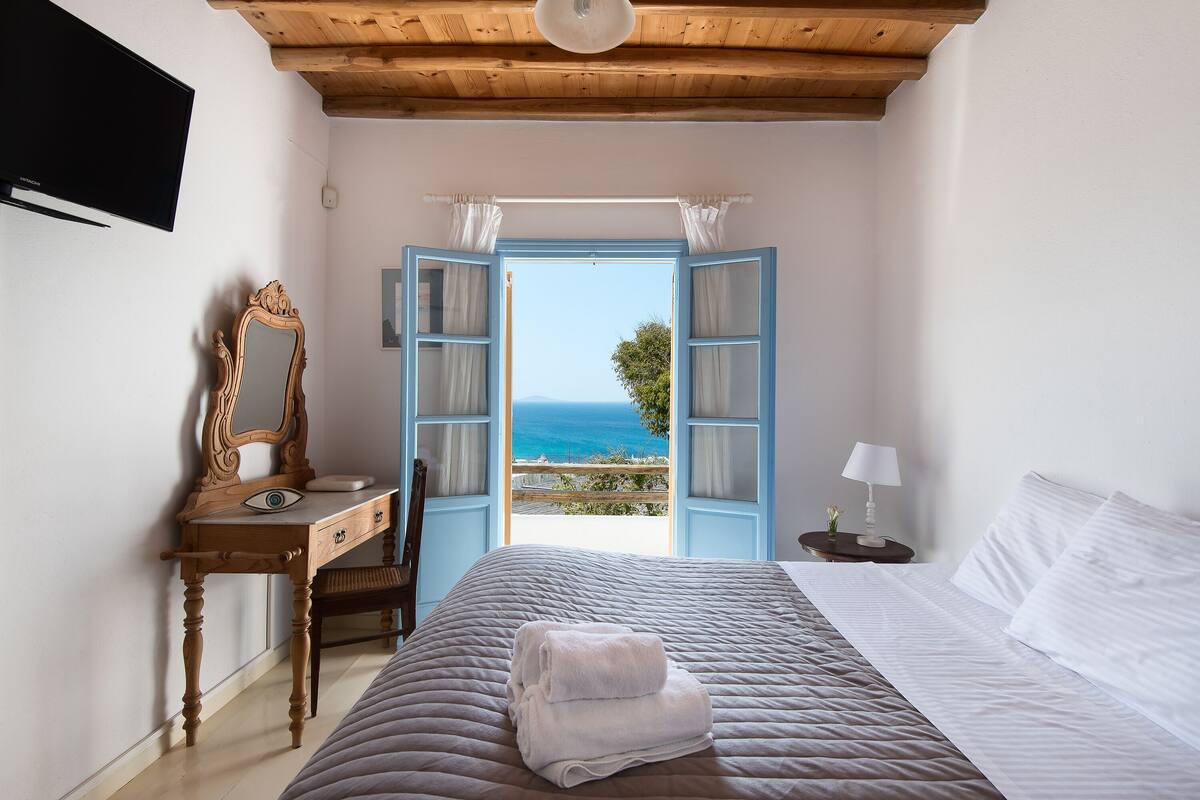 Un dormitorio con cama, un escritorio de madera con espejo, una mesita de noche con lámpara y una puerta abierta que da a un balcón con vistas al mar.