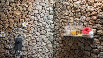 A stone wall with a metal hook, a shelf with bottles and a flower, and a towel.