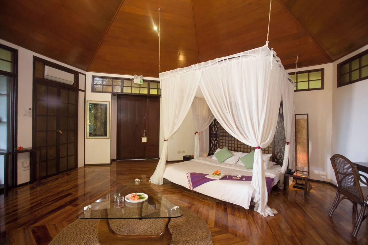 A room with a wooden ceiling, a bed with a canopy, a glass coffee table, and a chair.