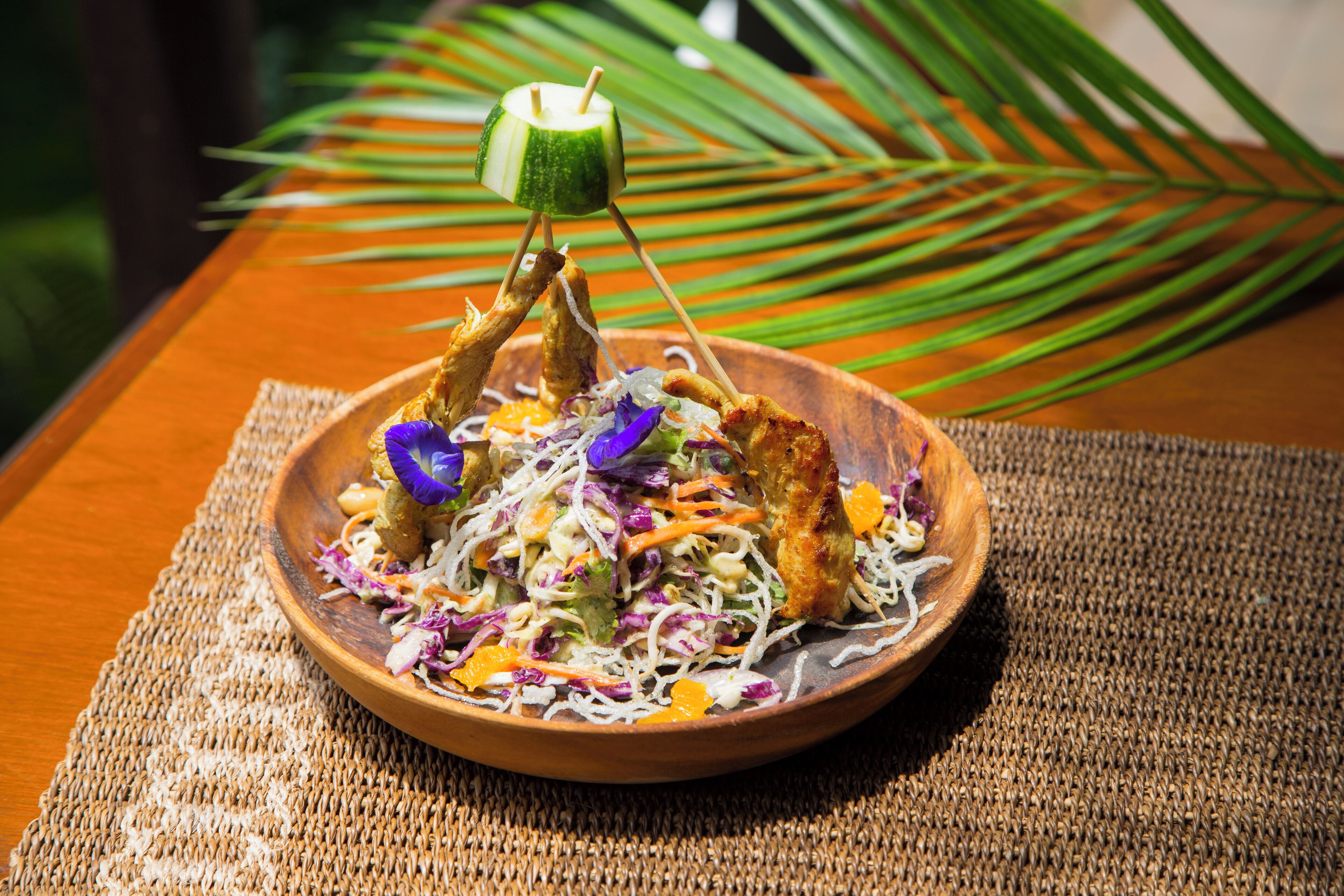 A wooden bowl filled with a salad and grilled meat, placed on a woven mat.