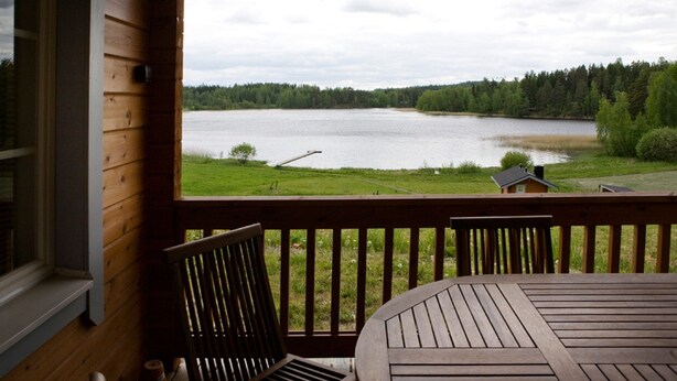 Una terraza de madera con una mesa y sillas da vista a un lago, rodeado de árboles y un área verde.