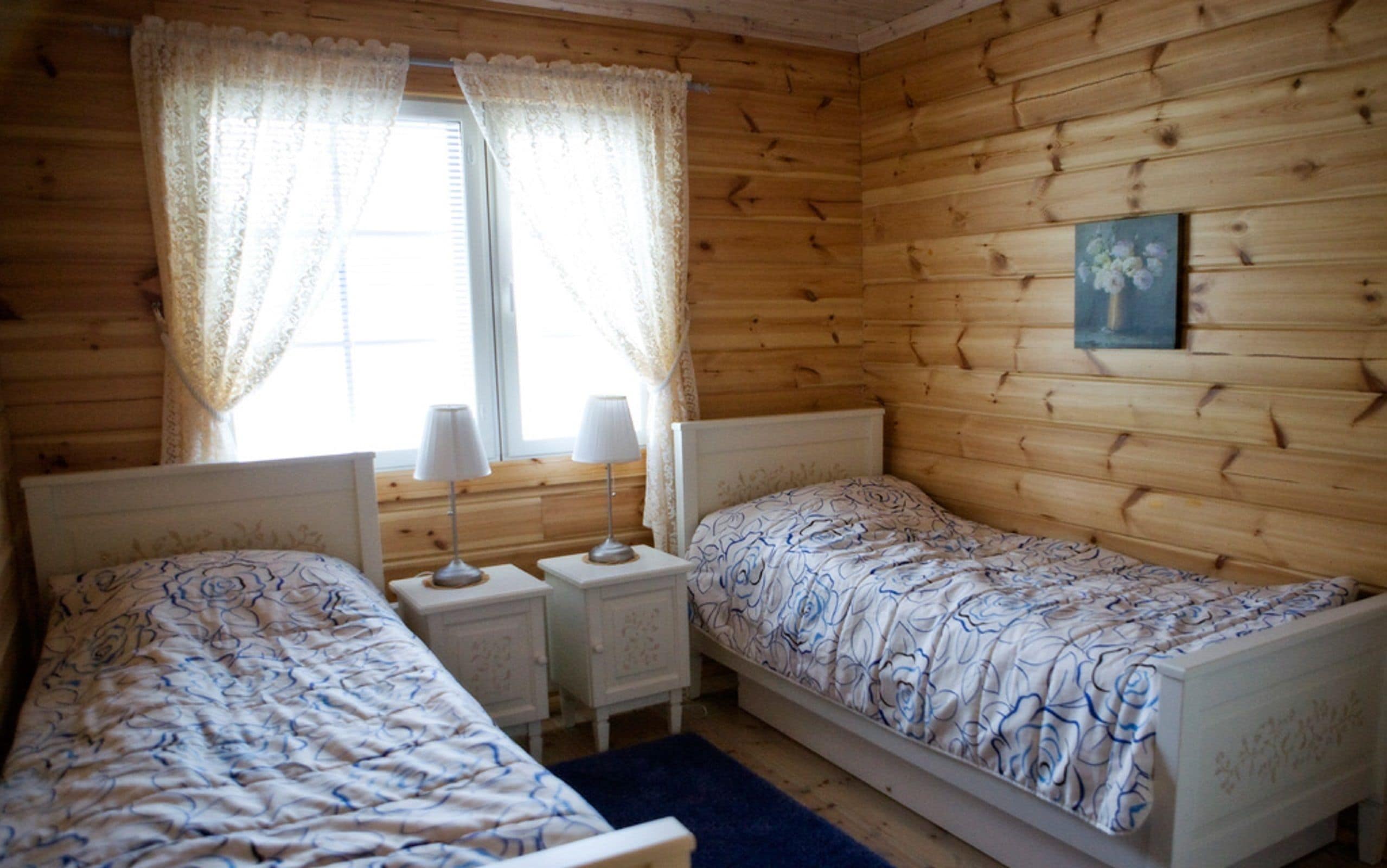 A wooden cabin room with two beds, a window with curtains, a bedside lamp, and a painting on the wall.