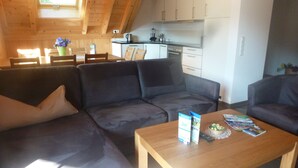 A cozy living area with a dark-colored sofa, a wooden coffee table with magazines and a bowl, and a kitchen in the background with white cabinets and a black countertop. - Rommelehof (Gutach)