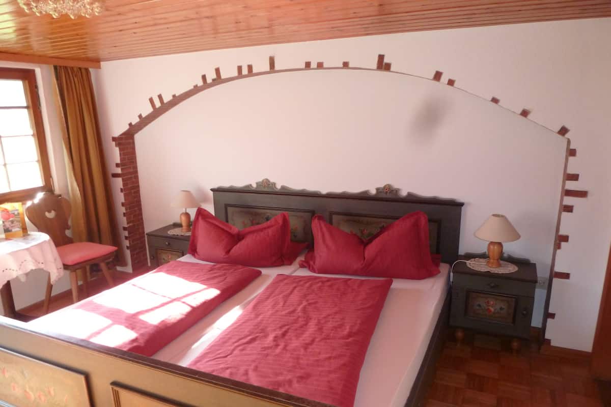 A bedroom with a wooden headboard, a bed with red bedding, two bedside tables with lamps, a chair, and a window with curtains.