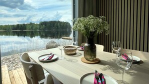 A dining table set with plates, cutlery, and wine glasses, overlooking a calm lake with a forested shoreline.
