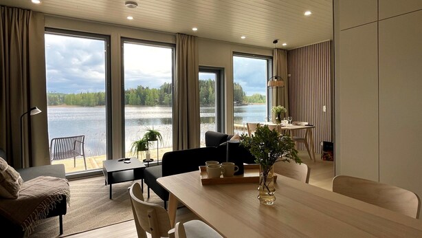 A modern living room with a large dining table, comfortable seating, and large windows overlooking a lake.