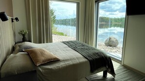 A bedroom with a bed, a nightstand, a lamp, a window with a view of a lake and forest, and a mounted TV.