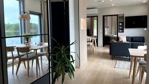 A modern hotel room with a dining area, a living area with a sofa and TV, and a large window overlooking a lake.