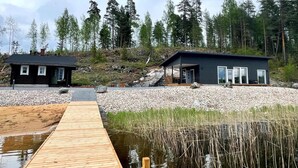 A modern cabin with a wooden dock extending into a calm body of water, surrounded by a forested landscape.