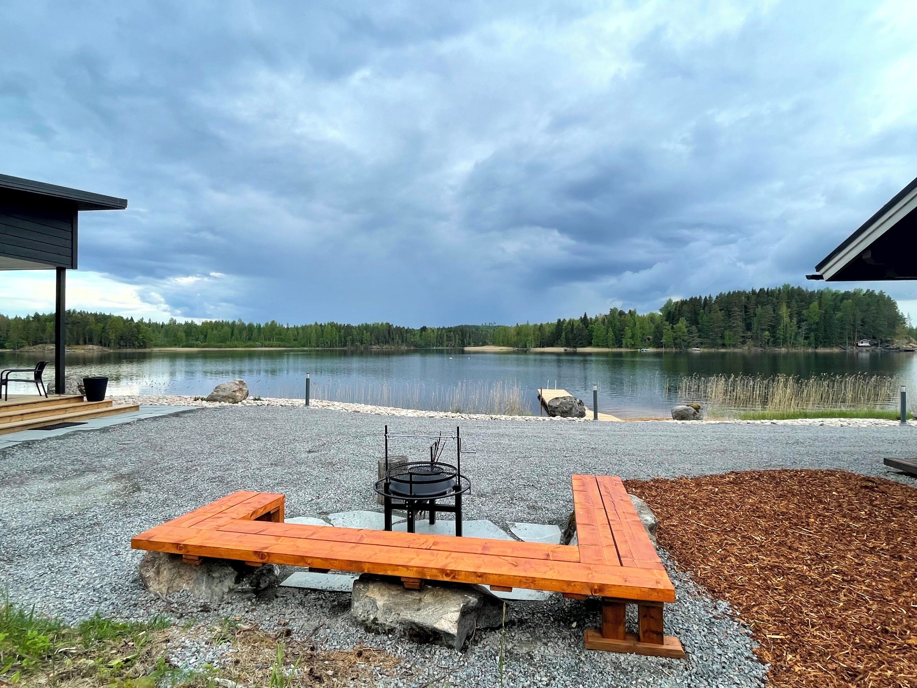 Een terras aan het meer met een houten tafel en stoelen, een barbecue en uitzicht op een bebost meer.