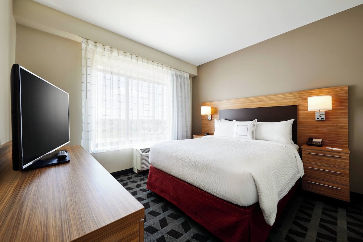 Une chambre d’hôtel avec un grand lit, un téléviseur posé sur un bureau en bois et une fenêtre ornée de rideaux légers.