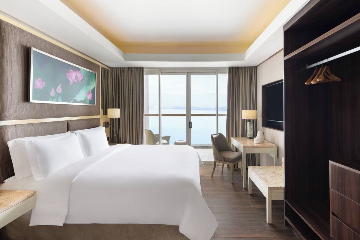 Ein Hotelzimmer mit einem großen Bett, einem Schreibtisch, einem Sessel, einem Fernseher und Blick auf das Wasser.