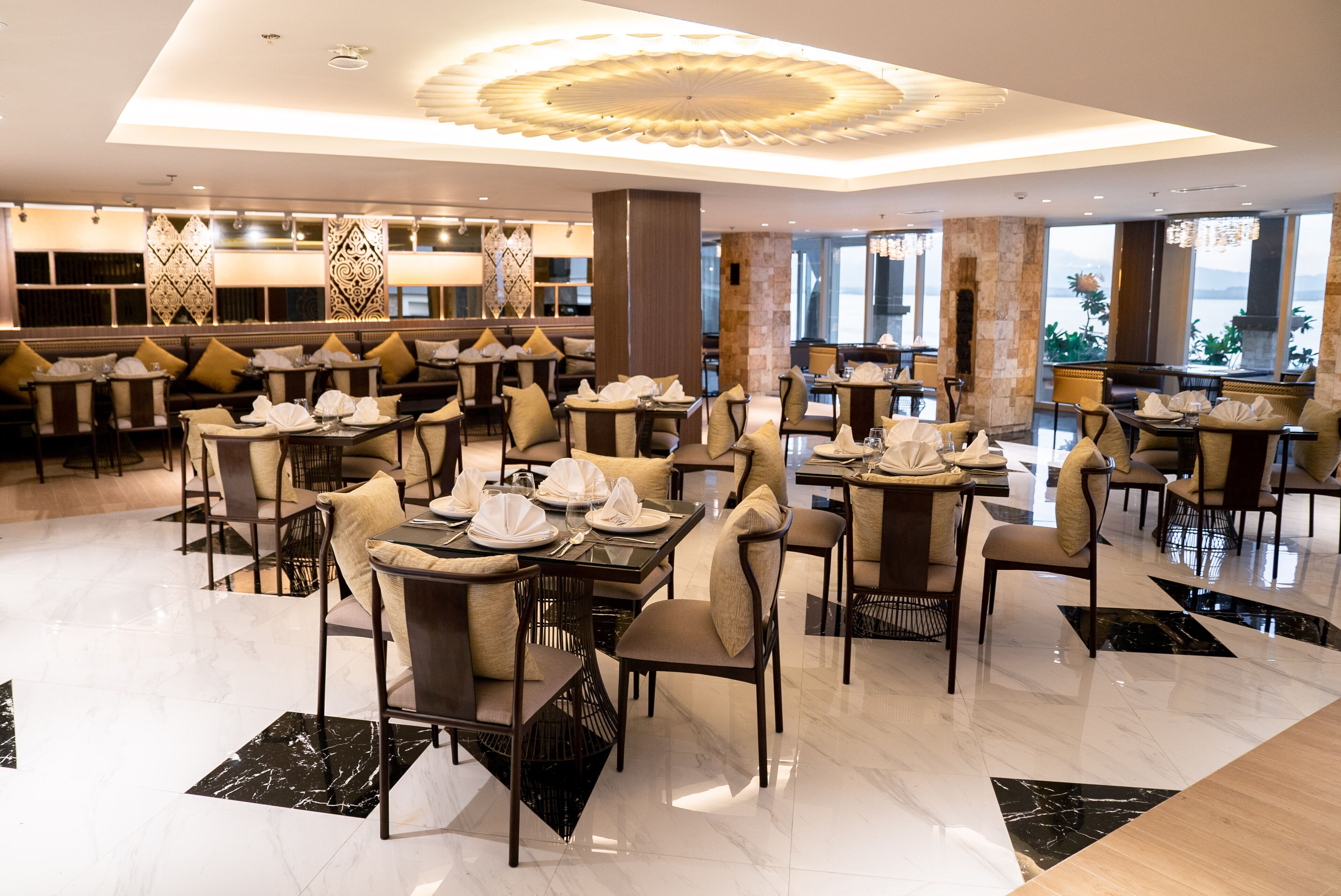 A spacious dining area with multiple tables and chairs, a decorative ceiling, and large windows offering a view of the outdoors.