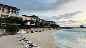 Una playa con sillas de descanso blancas y sombrillas, un muro de piedra y un edificio con balcones con vistas al océano.