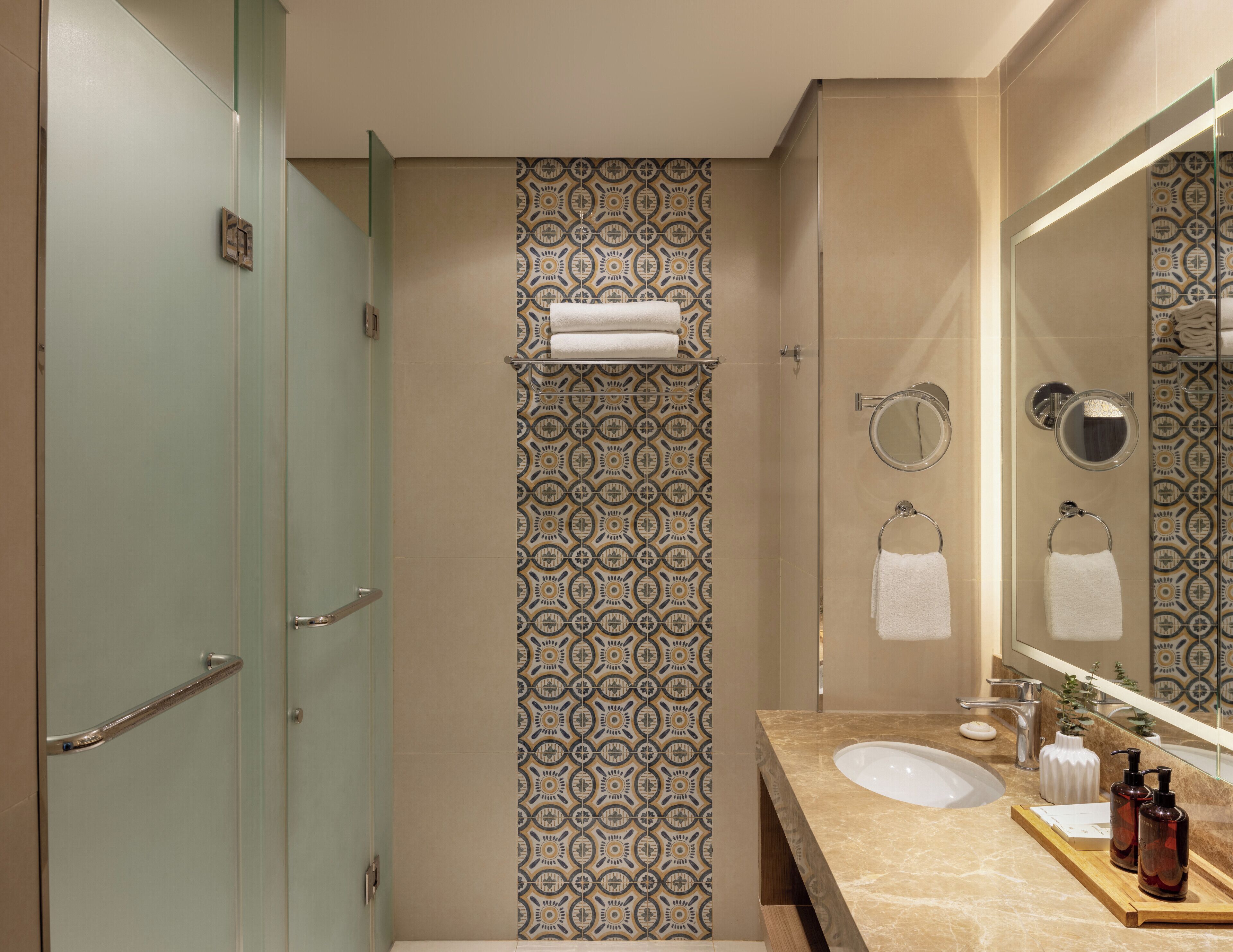 A modern bathroom with a glass shower enclosure, patterned wall tiles, a vanity with a sink, and a large mirror.